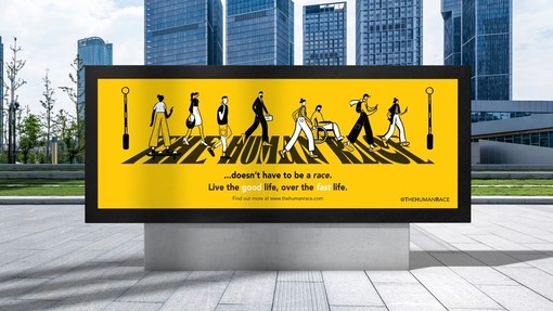 A virtual reality billboard in a clean cityscape with glass skyscrapers in the background; the yellow billboard shows diverse stylized figures in black walking across a zebra crossing; instead of zebra stripes, text under the feet of the characters reads The Human Race.