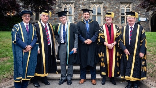 Saif Dai Rogers gyda Rhys Dart ac uwch aelodau eraill y Brifysgol tu allan i’r Hen Adeilad ar Gampws Caerfyrddin; maent i gyd yn gwisgo gwisg academaidd.