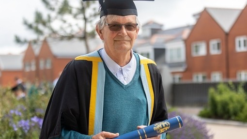 Martin Parr yn gwisgo gŵn academaidd du, cwfl glas a melyn, a het academaidd, ac yn gafael sgrôl ag arfbais gilt PCYDDS arno.