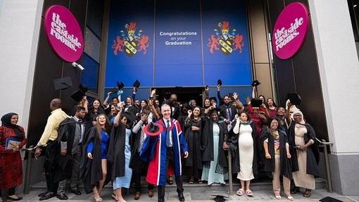 A group of about thirty graduates wave, laugh and smile as they leave the graduation ceremony. At the front of the group, the Dean of Birmingham Campus wearing blue and red robes holds out his hat and smiles.