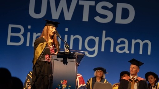 Wearing an academic gown and hat, Taya Gibbons stands at a microphone on stage.