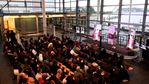 Mae siaradwr yn annerch cynulleidfa o lwyfan ym mynedfa’r Senedd; gellir gweld Bae Caerdydd a Phenarth trwy’r waliau gwydr tryloyw tu ôl iddi.