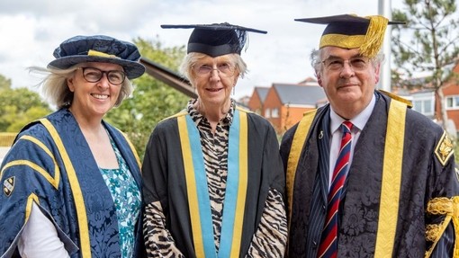 Gan gwisgo gwn ddu a chap academaidd, mae Pam Berry yn sefyll rhwng Jane O’Rourke a Medwin Hughes, y ddau mewn gynau ffurfiol.