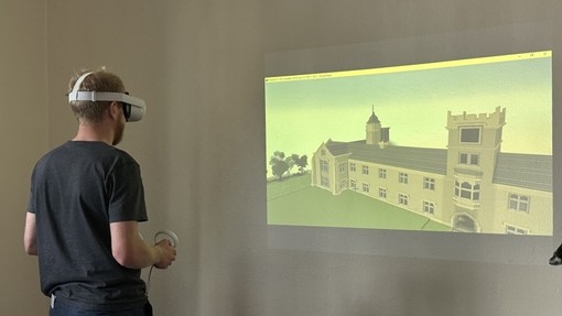 Myfyriwr yn gwisgo penset VR; mae tafluniad o Goleg Dewi Sant yn Llambed yn llenwi rhan o’r wal o’i flaen.