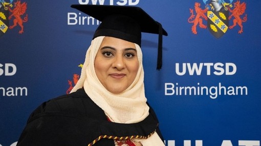 Umbereen Naeem yn gwenu at y camera ac yn wisgo cap a gŵn ar gyfer ei graddio.