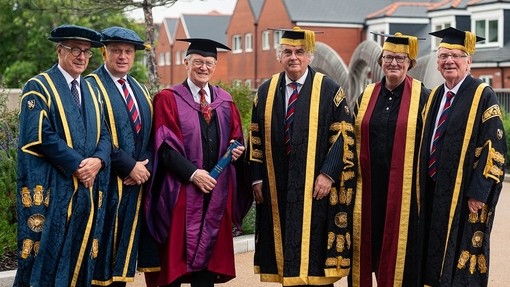 Y Gwir Anrhydeddus Alun Michael mewn gynau doethurol ysgarlad a phiws yn sefyll rhwng uwch aelodau’r Brifysgol.