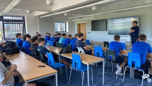 Ystafell ddosbarth yn llawn myfyrwyr gwrywaidd mewn crysau t glas; mae aelod o staff yn siarad ac mae sgrin fawr yn dangos y geiriau – The Rugby Academy.