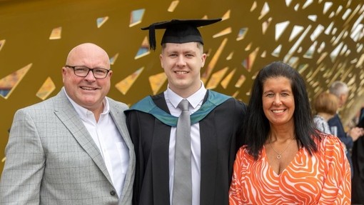 Yn ei ddillad graddio, mae Jac Elsey yn sefyll gyda’i freichiau o gwmpas ei rieni.