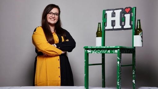 Kira Roberts yn gwenu wrth ymyl ei chynnyrch, cadair Heineken werdd gyda photel Heineken wag yn sownd wrth bob ochr a’r gair Hi mewn llythrennau mawr wedi’i osod ar y gefnell; mae symbol calon goch wedi cymryd lle’r smotyn ar ben y llythyren i.