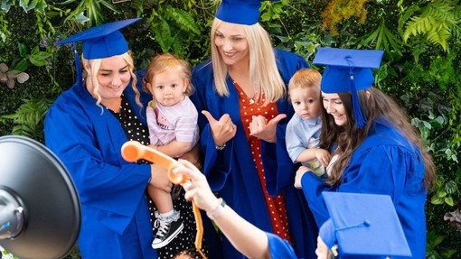 Three young women, two holding babies, smile while wearing blue robes and mortarboards.