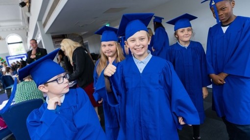 Children wearing blue robes and hats; a boy at the front of the group gives a thumbs-up to the camera.