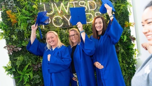 Three women in blue robes raise their mortarboards in front of a green wall of plants.