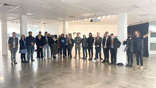 Cystadleuwyr rownd derfynol Cystadleuaeth Sgiliau Ffotograffiaeth Cymru yn sefyll yn Stiwdio Griffith; mae lluniau yn cael eu harddangos ar y waliau.