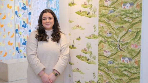 Ellie Jones yn sefyll gyda balchder wrth ymyl samplau o’i gwaith; mae’r samplau papur wal yn dangos golygfeydd o gefn gwlad.