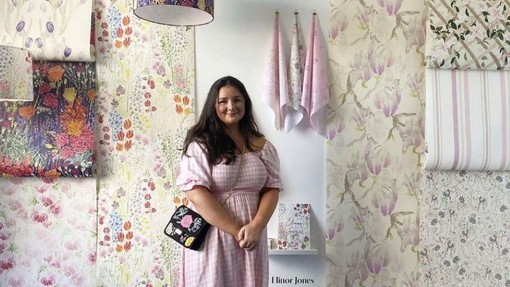 Ellie Jones yn sefyll gyda balchder wrth ymyl samplau o’i gwaith; mae’r samplau papur wal a thecstilau yn dangos patrymau blodeuog mewn lliwiau pastel a llachar.