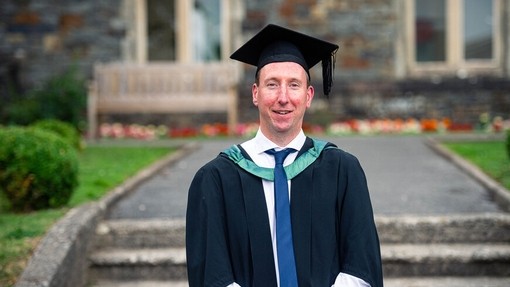 Gareth Howard mewn gŵn a het academaidd yn sefyll o flaen yr Hen Adeilad, Caerfyrddin.