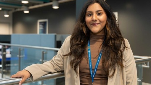 Amina Meah yn pwyso ei llaw ar falwstrad gwydr yn adeilad IQ.