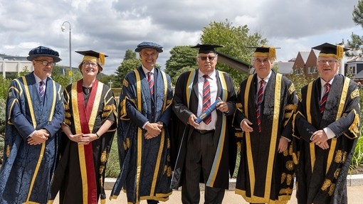 Tony Ball yn dal sgrôl glas a gilt ac yn gwisgo gŵn academaidd gydag uwch aelodau’r Brifysgol y naill ochr iddo.