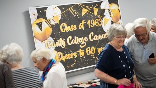 Friends look at photos and documents spread out on tables and talk; a banner on the wall reads – Class of 1963 Trinity College Carmarthen – Cheers to 60 years.