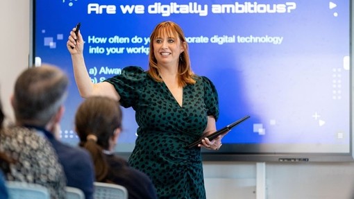 Siaradwraig yn nigwyddiad yr IDNS ar Dechnolegau’r Dyfodol, sef ‘Matter 2023’ yn sefyll o flaen sgrin sy’n dweud ‘are you digitally ambitious?’ ac yn gofyn i’r gynulleidfa ymateb