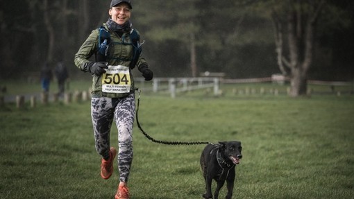 A female participant running in gloves and a jacket beside a small dog keeping pace with her.