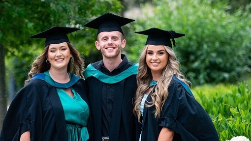 A young man and two young women in academic robes, hoods and hats smile for the camera.