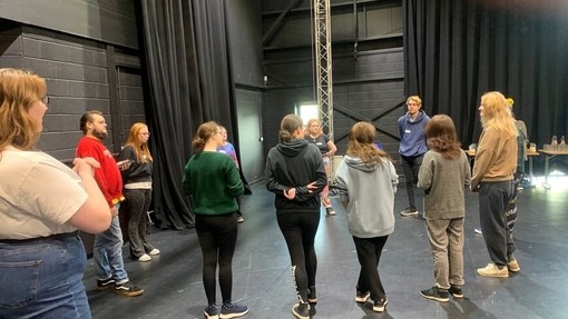 Grŵp o bobl yn cymryd rhan mewn gweithdy Theatr fel Arfer Cynhwysol ac yn gwrando ar yr hyfforddwr
