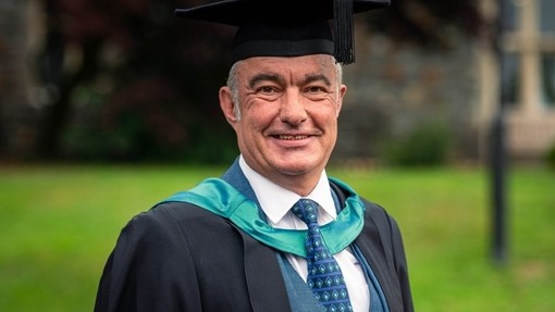 Gan wisgo gwisg academaidd, mae Ken Dicks yn gwenu tuag at y camera.