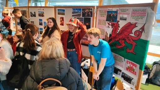 Grŵp o fyfyrwyr wrth y stondin sy’n cynrychioli Cymru a PCYDDS yn ystod wythnos Ryngwladol Coleg Prifysgol Volda; mae’r stondin wedi’i haddurno â lluniau o Gymru a baner Cymru ac mae myfyriwr yn gwisgo het fwced.