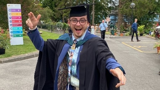 Lewis Thornton yn gwenu ac yn mwynhau mas draw yn ei ŵn graddio.