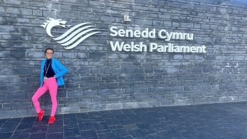 Tilley Rees, yn gwisgo legins pinc llachar a blaser glas, yn pwyso yn erbyn wal lechi gyda llythrennau gwyn sy’n dweud: Senedd Cymru – Welsh Parliament.