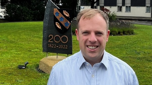 Dan Priddy yn edrych i gyfeiriad y camera; ar y glaswellt tu ôl iddo mae cerflun carreg yn coffa daucanmlwyddiant y Brifysgol.