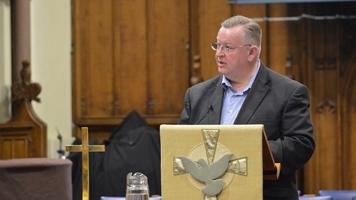 Yr Athro Williams Gibson wrth ddarllenfa wedi’i haddurno â lliain ag arno golomen yn hedfan o flaen croes.
