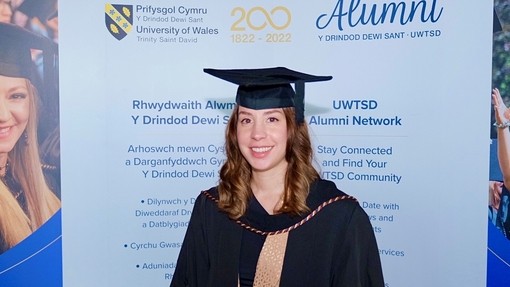 Mewn gwisg academaidd, gwena Yolanda Rendón-Guerrero i gyfeiriad y camera; mae hi’n sefyll o flaen arwydd yn hyrwyddo Rhwydwaith Cyn-fyfyrwyr PCYDDS.