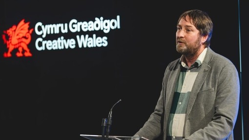 Dyn yn siarad tu ôl i bodiwm; mae draig goch nesaf at y geiriau Cymru Greadigol yn Gymraeg a Saesneg i’w gweld ar sgrin y tu ôl iddo.