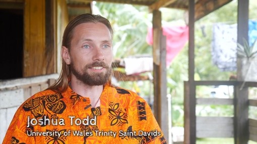 Joshua Todd, yn gwisgo crys oren â motiffau glöynnod byw du, yn eistedd ar feranda tŷ yn Ffiji.