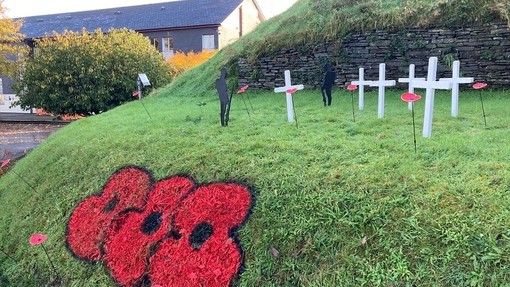 Twmpath gwyrdd wedi’i addurno â chroesau gwyn, silwetau du o filwyr, a phabïau coch mawr wedi’u paentio â chwistrell ar y glaswellt.