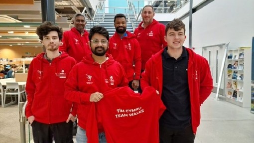 Yn sefyll ar y grisiau canolog yn Adeilad IQ, mae chwe myfyriwr yn cynnwys Rohan Chavan a David Ntino yn gwisgo hwdis coch ac yn dal crys chwys coch Tîm Cymru/ Team Wales.