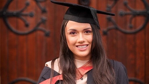 Mae Faith Over yn gwenu yn ei gynau graddio.