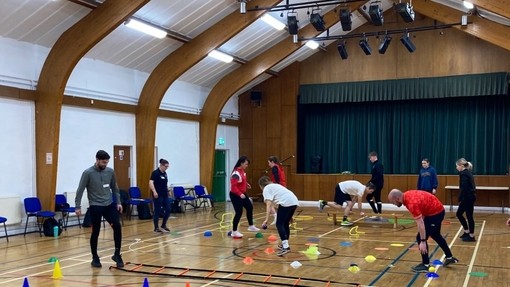 Pobl ifanc yn eu harddegau yn symud rhwng conau plastig mewn campfa ysgol.