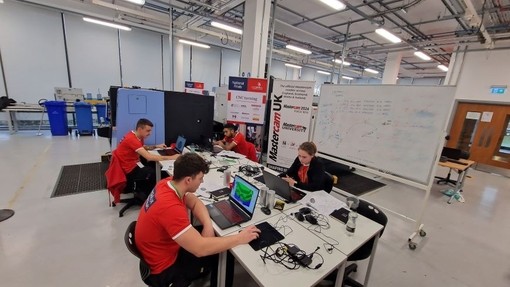 In a large white hanger, three young men and one young woman sit at a table focusing intently on their laptops.