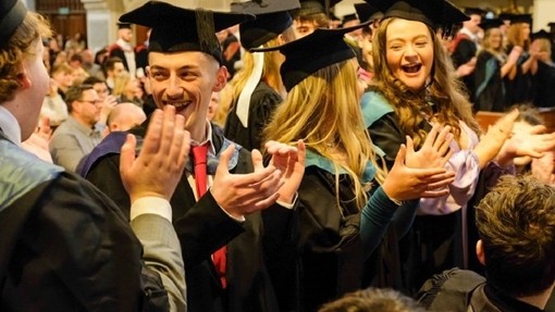 Graddedigion mewn gynau’n gwenu a chlapio yn ystod y seremoni.