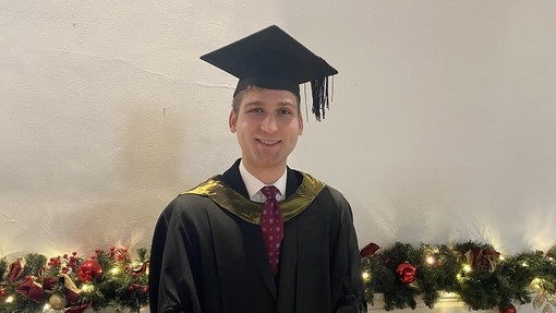 Yn gwisgo gŵn a het academaidd, mae Brandon Gorman yn sefyll o flaen silff ben tân ag arno arlant Nadolig.