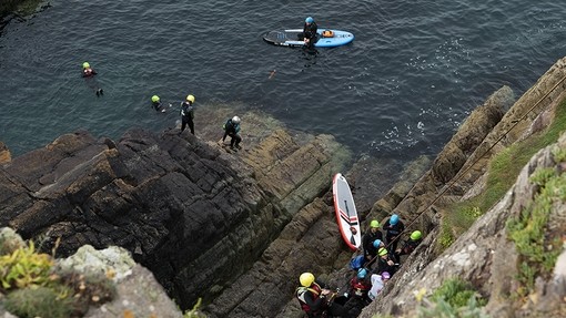 A group in wetsuits and helmets climb from the sea onto a rock before they begin scaling a sea cliff.