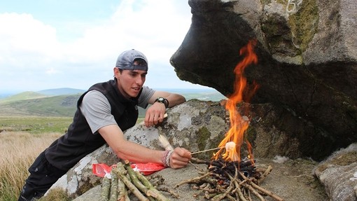 A young man toasts a marshmallow in a small wood fire on a boulder.