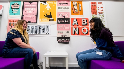 Two laughing female students sit opposite each other on purple couches.