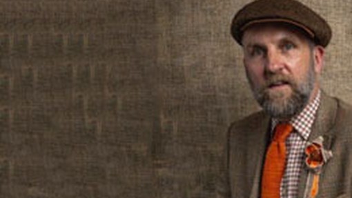 Mae Marc Rees yn gwisgo tei oren a blodyn brethyn patrymog oren dros boced ei frest; mae cysgod brown golau ei flaser gwlân yn cyfateb i'r wal y tu ôl iddo; mae'n gwisgo cap fflat brown traddodiadol.