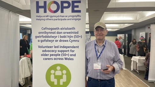 Dennis Warwick yn sefyll wrth ochr baner godi â’r geiriau: Hope, cefnogaeth eiriolaeth annibynnol dan arweiniad gwirfoddolwyr i bobl hŷn (50+) a gofalwyr ar draws Cymru.