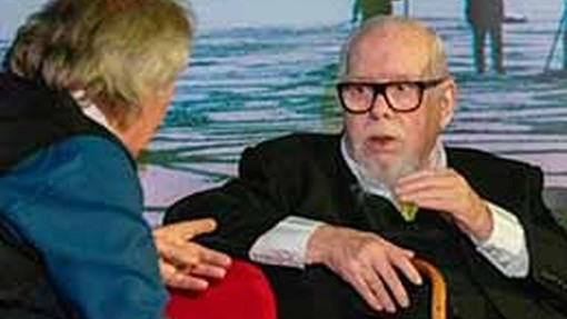 Syr Peter Blake yn eistedd mewn cadair yn ystod cyfweliad ac yn codi llaw i bwysleisio.