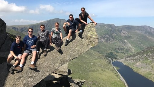 A group of students sit and smile on a rocky outcrop that juts out above a deep valley.